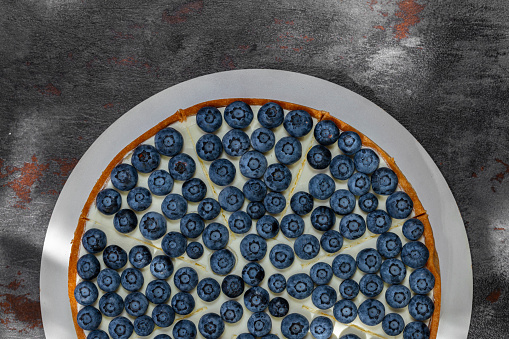 Shortcrust pastry pie with creamy vanilla cream and blueberries. The pie stands on a light stand on a gray background with bronze inserts.