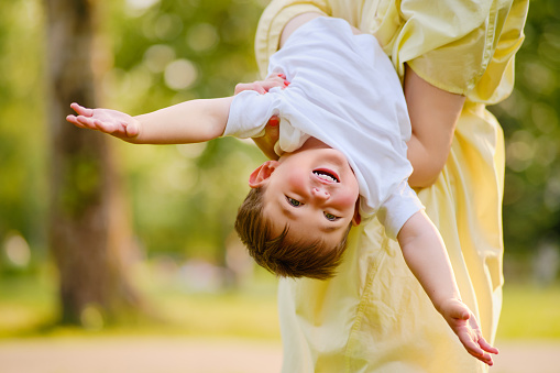 A child upside down in the arms of his mother on a walk in nature. Toddler baby and mom play on the green grass in the summer park. Kid aged about two years (one year eleven months)