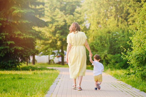 A child takes steps holding a mother woman by the hand along a path in nature. Happy baby with mom on a walk in the summer park. Kid aged about two years (one year eleven months)