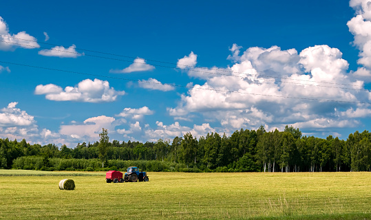 Agricultural countryside landscape with tractor and haystacks, forest and clouds above
