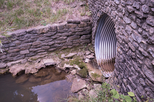 Corrugated metal sewage drainpipe draining water into creek with stone walls