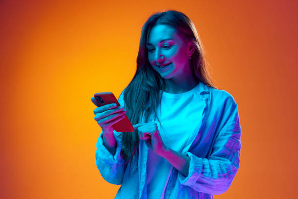 istock Smiling young woman typing text message on mobile phone against gradient orange studio background in neon light. Online communication 1503354271