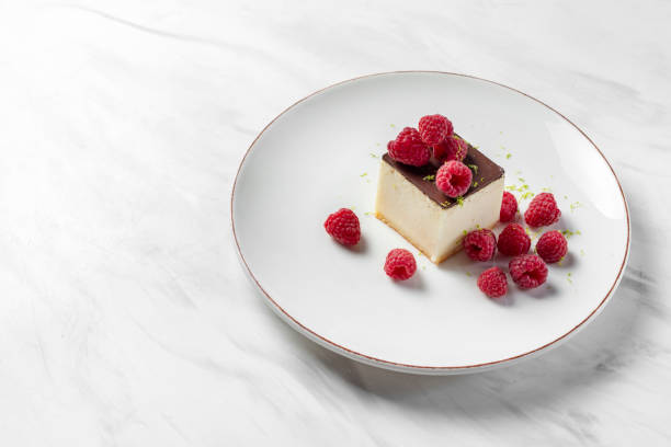 Dessert cheesecake on a nut crust with chocolate icing. Raspberries and lime zest lie on top and next to it. The dessert lies on a light ceramic plate on a marble surface.