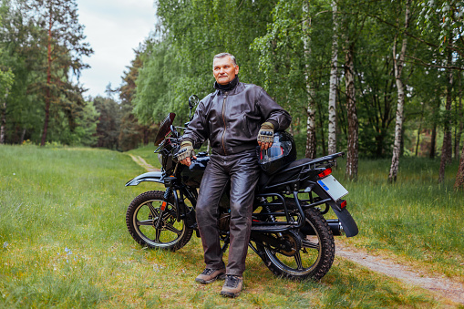 Elderly biker leaning on motobike in summer forest. Man in leather clothes relaxing enjoying nature after ride