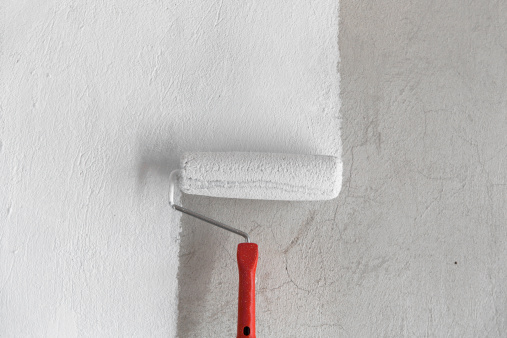 The image contains a painting roller with white paint. The roller is in the center and devides the frame into two parts: painted and non painted. The wall is a rough coat. The colors are neutral. 