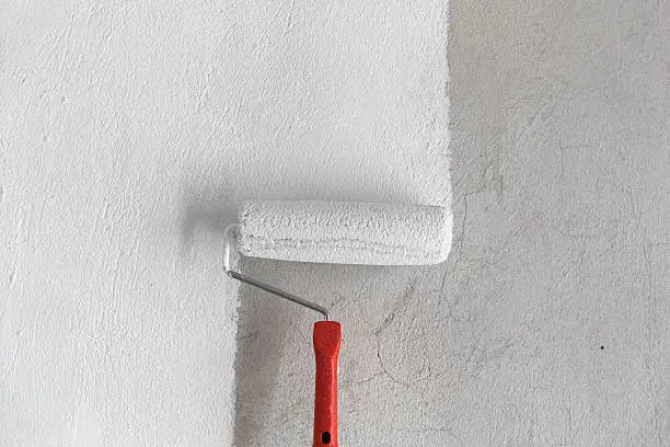 The image contains a painting roller with white paint. The roller is in the center and devides the frame into two parts: painted and non painted. The wall is a rough coat. The colors are neutral. 