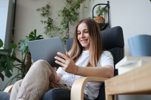 Young adult women holding coffee cup while looking away. Female is sitting on sofa at home. She is in casuals. Shot of a happy young women using his tablet while relaxing on the couch at home