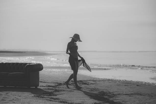 Young woman pose in fashionable clothes on the sand beach. Beautiful sea landscape on background. Brunette model, tall, tan and slim. Summer, sunset. Black and white picture.