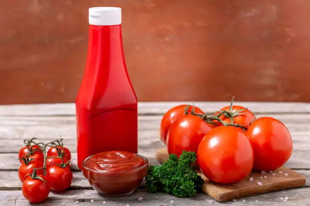 Plastic bottle and glass bowl of ketchup or tomato sauce, spices and fresh tomatoes on wooden table. Selective focus.