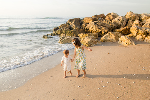 4-Year-Old Toddler Girl Holding Her 1-Year-Old Brothers Hand While Walking on the Beach, Enjoying Time Together During a Golden Sunrise in Palm Beach, FL in the Summer of 2023.
