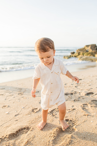 A 1-Year-Old Cuban-American Baby Boy Walking on a Sandy Beach Wearing a Cream-Colored Linen Romper While Exploring the Beach During a Golden Sunrise on Palm Beach, FL in the Summer of 2023