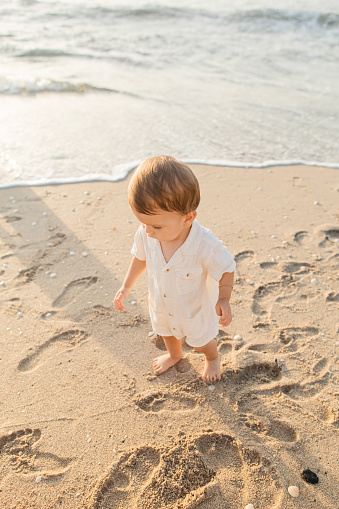 A 1-Year-Old Cuban-American Baby Boy Walking on a Sandy Beach Wearing a Cream-Colored Linen Romper While Exploring the Beach During a Golden Sunrise on Palm Beach, FL in the Summer of 2023