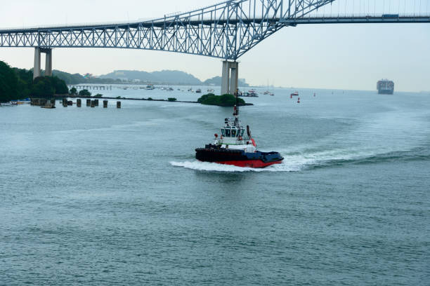 panama canal tug boat passes under puente de las americas on the panama canal. freighter in the background - panama canal panama global finance container ship imagens e fotografias de stock