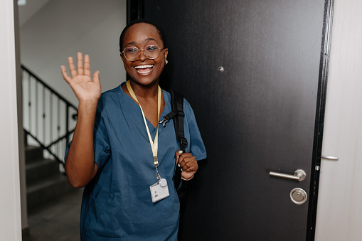 A young African American female nurse is cheerfully smiling and waving while entering her apartment.