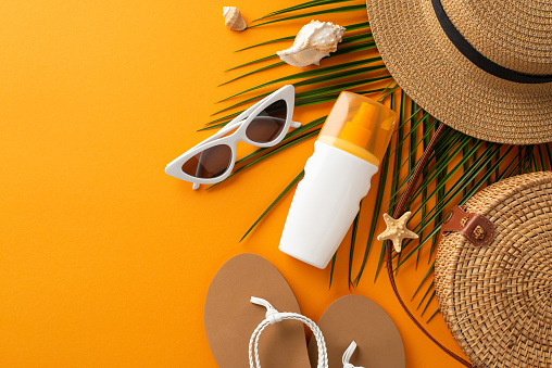 Ultraviolet protection on the beach concept. Top view photo of shells and palm leaves, straw hat, wicker bag and flip-flops and sunscreen on orange isolated background with copyspace
