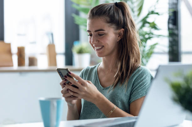 istock Concentrated young business woman using her mobile phone while working with laptop in the kitchen at home 1503443372