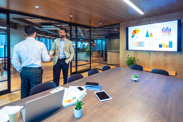 Business men handshake in a board room. There is a whiteboard presentation with finance data charts