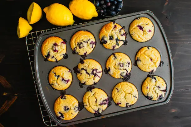 Freshly baked blueberry muffins in a muffin tin with lemons and blueberries on the side