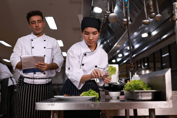 istock Chef teaching how to cook Chef teaching the student how to prepare wok dish, Preparing vegetable on a plate. A female Chef in the kitchen provides cooking training to her students. Male chef trainees happily look and cook together. Chefs wearing a cooking 1503491560