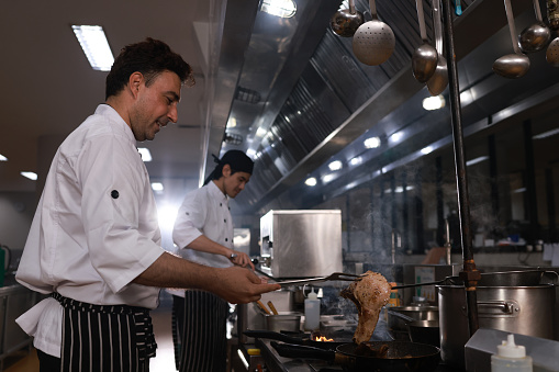 Chef's hands keep wok with fire. Closeup chef hands cook food with fire. Chef man burns food in a professional kitchen. Shot of two cooks preparing food in the kitchen. Cooks prepare meals on the stove in the kitchen of the restaurant or hotel. The fire in the kitchen.