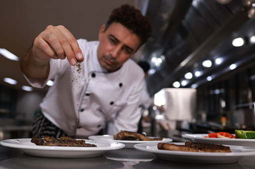 Male cooks are cooking with salt or spice hand sprinkle on steak and arrange to decorate a plate with team cooks. Chefs in hotel or restaurant kitchen cooking, Multiracial team of cooks cooking by kitchen stove in the restaurant, chefs cooking.