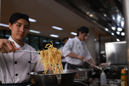 Young beautiful Asian woman chefs enjoy cooking healthy food with tong spaghetti or noodle  in cooking blow  on the stove in the kitchen at restaurants with  chef team