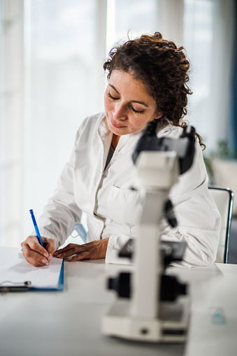 Selective focus shot of mid adult female laboratory technician writing results on the clipboard while analyzing scientific samples under a microscope when conducting research at the laboratory.