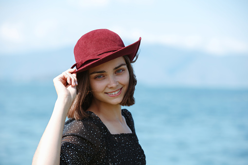 Woman smiling and touching hat while standing against blue sea and looking at camera enjoying summer holiday