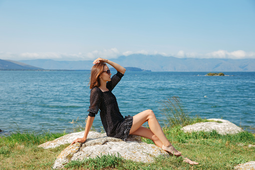 Carefree woman in short summer dress sitting on rock on beach and enjoying vacation near sea