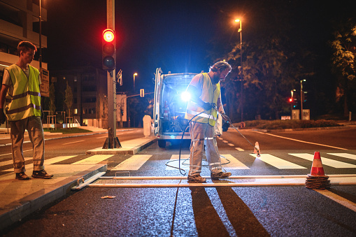 Road maintenance crew wearing reflective vests spray painting a new layer of paint on a crosswalk.