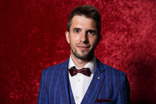 Content bearded handsome showman in elegant suit and red bow tie looking calmly at camera on red background in studio