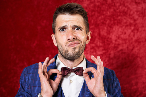 Hilarious male presenter in suit and bow tie having fun and looking at camera in studio on red background