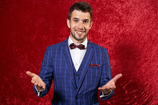 Cheerful showman in elegant blue suit and bow tie showing welcome gesture with hands and looking at camera with smile against red background in studio