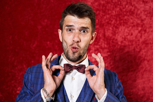 Positive showman in suit touching bow tie and making funny face while grimacing and looking at camera on red background during show