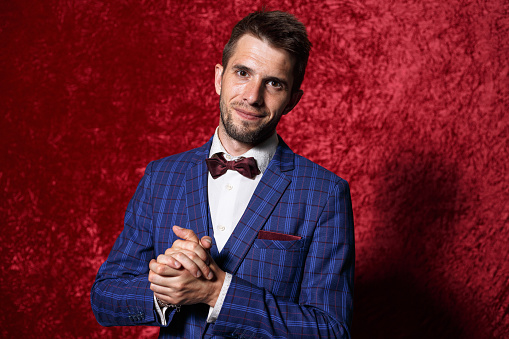 Content adult showman wearing blue suit and bow tie looking at camera with smile on red background