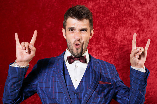Excited man in blue suit showing rock gesture or horn sign and looking at camera against red background in studio