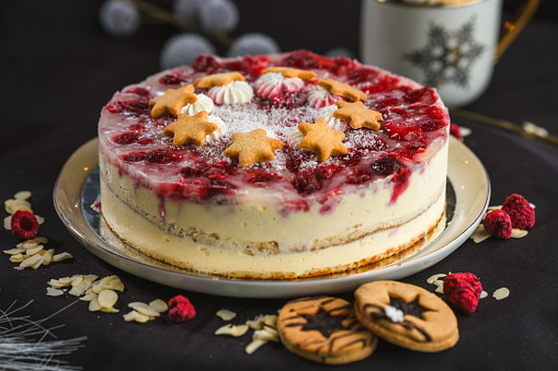 High angle view of a whole   raspberry mascarpone cake presented on a tray, in front of a black studio background, raspberries and almond flakes scattered in the foreground, cookies next to the plate. Studio shot, full length image.