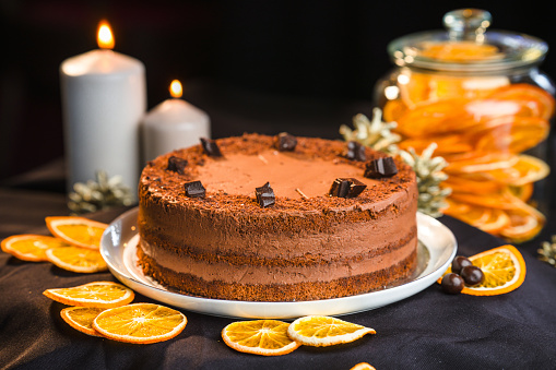 Whole, round chocolate cake on a ceramic platter, beautifully arranged on a black studio background with dried lemon slices scattered around and a jar full of them, lit candles in the blurred background. Studio shot, full length image. Side view.