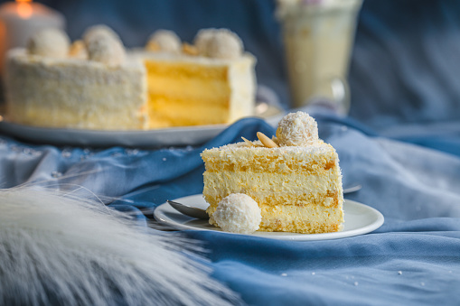 Vegan coconut and almond cake, displayed on a blue studio background, focus on a big slice of the same cake in  foreground. Side view, studio shot, full length image.