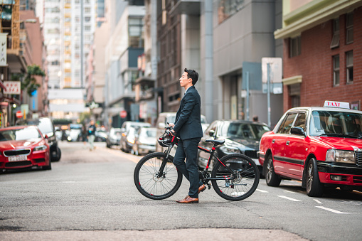 Side view of a young male Asian corporate employee crossing the street with a bicycle. He is walking with the bicycle next to him.
