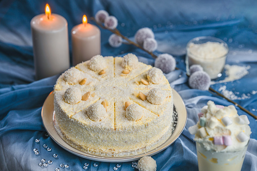 Professionally made  coconut and almond cake, displayed on a blue sheet, arranged with glass beads, lit candles, coconut flakes in a cup and plant decor in the background, focus on a tall glass of milk shake with marshmallows next to the cake.  Studio shot, full length image, high angle view.