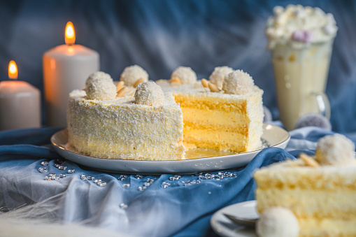 Gluten free, surprise birthday  cake, decorated with white cream and coconut pralines, arranged  with lit candles and feathers, a tall glass of milk  shake with marshmallows in the blurred background.  A big chunk of the same cake visible in the blurred foreground. Side view, studio shot, full length image.