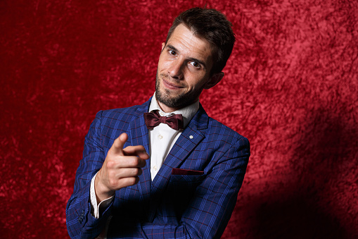 Showman in blue suit and bow tie pointing at camera and showing you gesture while standing against red background in studio