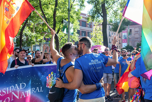 Madrid, Spain, 6th jul 2019. Two guys kissing while holding Spanish and rainbow flags during Gay Pride parade. The crowd enjoys.