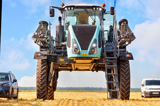 A high wheelbase industrial sprayer for the agricultural sector stands in comparison with passenger cars in a harvested field against a blue cloudy sky.