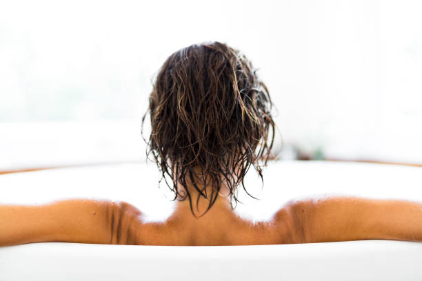 Point-of-view shot of a person soaking feet in a bathtub; chrome grab bars and mixer tap mounted on mosaic tiles suggest accessibility and home self-care.