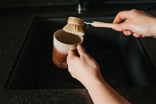 Mugs cleaning with a brush.Ecofriendly housekeeping and dishwashing. hands wash a ceramic mug with a wooden brush with natural bristles over a black sink .
