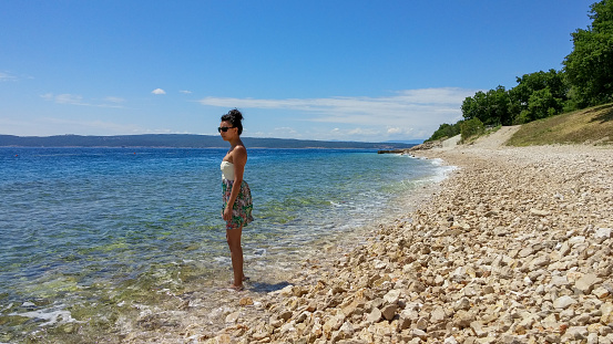 A tall woman stands in the waters of the Adriatic Sea in Croatia. A sunny day with beautiful blue water in the town of Novi Vinodolski.