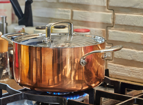 Copper saucepan with lid with dish is cooked over fire on gas stove. Steam rises above the pot.