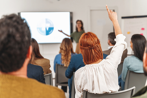 Businesswoman with red hair raising hand to ask question during a SWOT analysis presentation in large office room. Diverse audience, viewed from back.